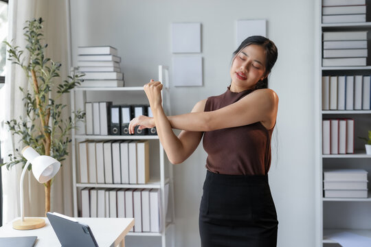 Young businesswoman stretching at her office desk, surrounded by typical items. Natural light highlights the background