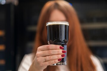 Close-up photo of a woman with red nail polish holding a glass of dark beer with a creamy head. The background is blurred, focusing on the beer and the woman's hand
