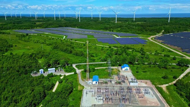 Solar panel farm and wind turbines supplying an electrical substation in Estonia's countryside by the seashore - aerial flyover