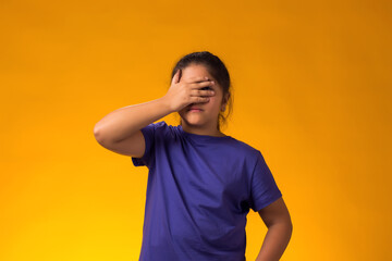 Upset kid girl covering face with palms and looking through fingers over yellow background. Emotions and bulling concept