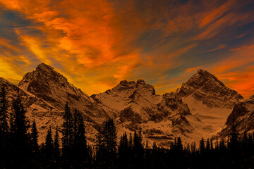 Dramatic colourful cloud formation above a snow-covered mountain range and silhouetted evergreen trees in the foreground, south of Canmore, Alberta; Alberta, Canada