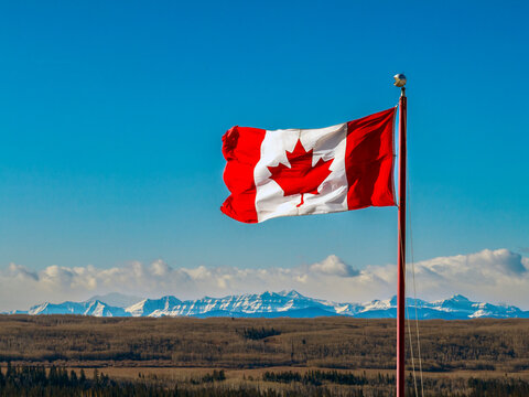 Close up of a Canadian flag blowing in the wind, with snow-covered mountain range, blue sky and clouds in the background; Calgary, Alberta, Canada