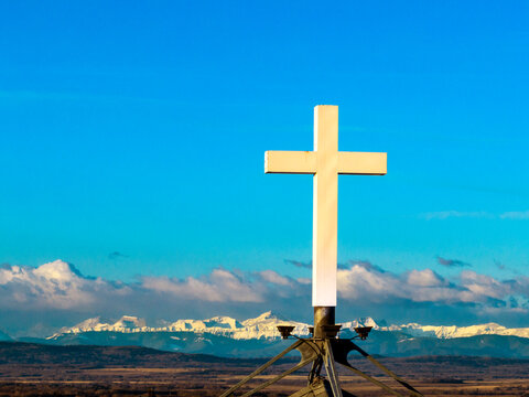 Close up of a church steeple's white cross with blue sky, clouds and snow covered mountain range in the background; Calgary, Alberta, Canada