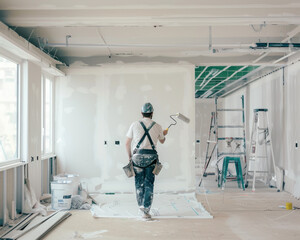 Professional studio photo of a worker using a paint roller on a white wall inside a new built house, under construction.