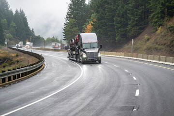 Naklejka premium Gray bonnet big rig semi truck transporting cars on car hauler semi trailer driving on the winding highway road with rain and fog