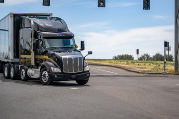 Shiny black big rig semi truck transporting cargo in dry van semi trailer turning on the crossroad...