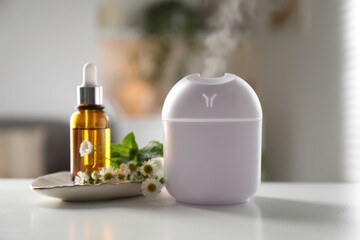 Essential oil diffuser, cosmetic product and chamomile flowers on white table at home