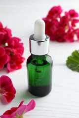 Bottle of geranium essential oil and beautiful flowers on white wooden table, closeup