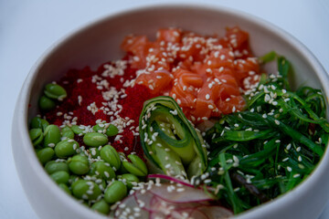 Close-up view of seafood poke bowl with salmon, rice, cucumber, radish, nori seaweed and edamame beans on white table in restaurant or sidewalk cafe. Soft focus. Copy space. Food theme.