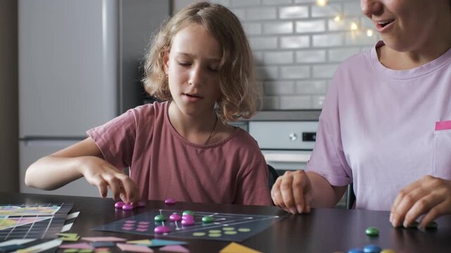 A young girl and her mother play a board game together in their kitchen, enjoying quality time and laughter, slow motion. Family lifestyle