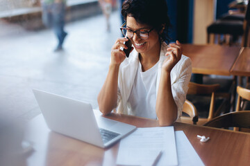 Smiling businesswoman engaged in a phone call during a remote working session at a cafe, focusing on a project with her laptop.