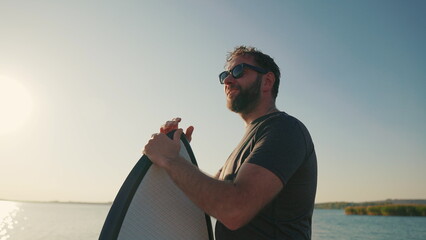 Close-up portrait of a man standing on a lake pier at sunset, leaning on a waterboard. Male surfer...