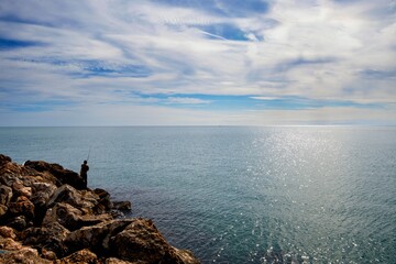 Lone fisherman standing on rocks by the sea under a partly cloudy sky on a sunny day