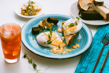 Eggs Benedict with fried toast and crouton on a turquoise plate and orange juice with ice on a white background. Selected focus