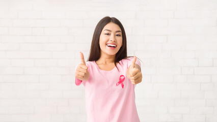 Cheerful Asian Girl In Pink Ribbon T-Shirt Gesturing Thumbs Up Supporting Cancer Awareness Campaign Standing Over White Background. Selective Focus