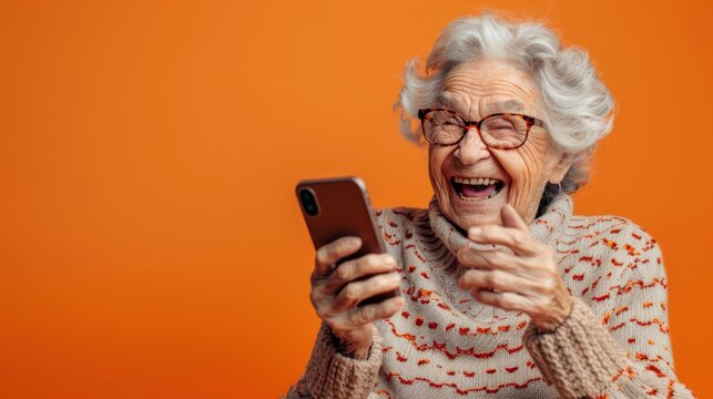 An elderly woman with gray hair is joyfully smiling and laughing while holding her phone against a vibrant orange background.