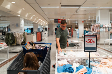 A man shopping for clothes in a store while his child sits in the cart. He examines a rack of discounted clothing. Male consumer, bargain hunter, frugal living with family 