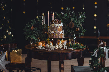 Elegant beautiful Christmas house decoration, stylish holiday decor. Homemade delicious sweet gingerbread house as centrepiece for family dinner. Fir tree branches, candles, stars, garlands