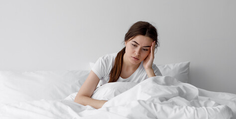 Sad european millennial female, worried, suffers from insomnia and headache. Lady sits on bed in...