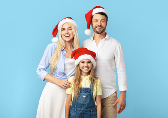 Obraz premium A young girl stands between her parents, all three smiling and wearing Santa hats. The family is standing in front of a solid blue background.