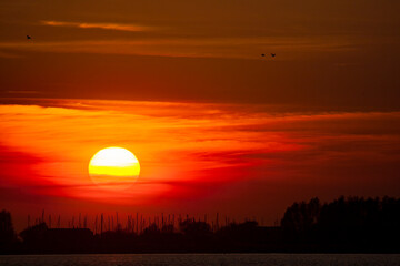 A vibrant red sunset over a wide river, with tree silhouettes and a marina visible on the horizon, creating a picturesque scene.