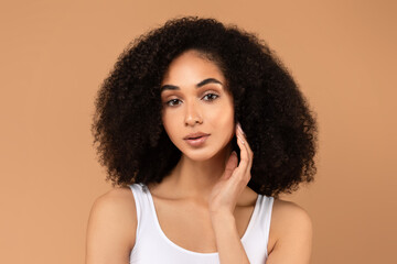 A young black woman with dark, curly hair is posing in a white tank top against a tan background. She has a hand resting on her cheek and is looking directly at the camera