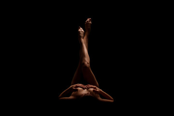 A woman lies on her back in a darkened room, her legs raised in the air with her toes pointed. The...