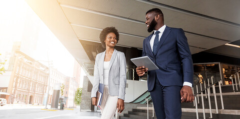 Two smiling african american colleagues discussing new project while going out of office building, copy space