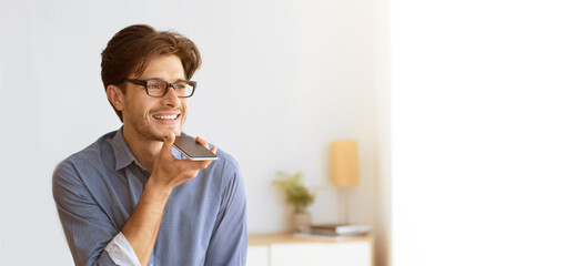 A man in a blue shirt and glasses smiles while using his smartphones voice assistant indoors with a lamp and plant in the background.