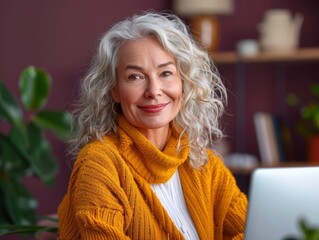 A smiling mature businesswoman with gray curly hair, wearing a yellow sweater, sitting in an office environment while working on a computer.