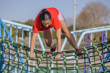 Latin girl in red t-shirt climbing a net in a public park.