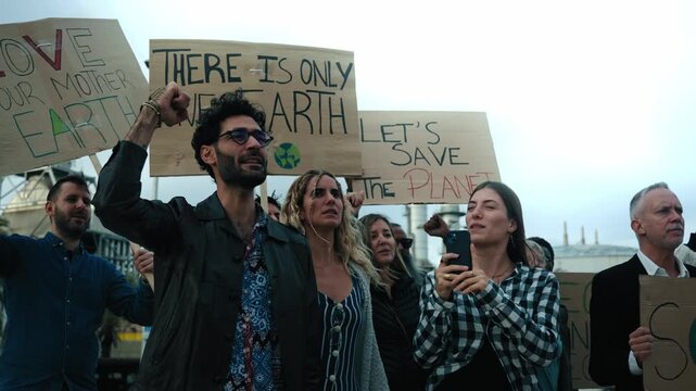 Group of diverse activists people at a demonstration against pollution and factories angrily shouting a slogan while raising their fists and holding several banners with messages about global warming