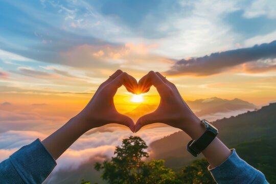 Hands forming a heart shape at sunset over a mountainous landscape - Powered by Adobe