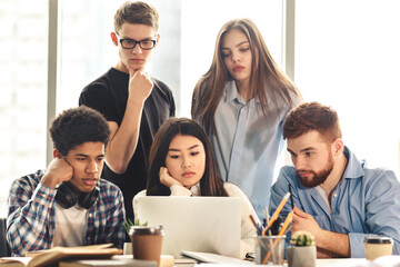 Fototapeta premium A group of five young professionals is gathered around a laptop, collaborating intently on a project. Some appearing pensive and others focused, they work together in a sunlit office setting