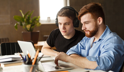 Two guys are seated at a desk in an office environment, focused on a laptop screen. They appear to be engaged in collaborative work, possibly discussing or analyzing data