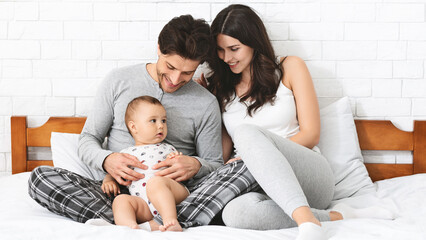 A happy family sits on a white bed in a bedroom. The father is holding their baby, and the mother...