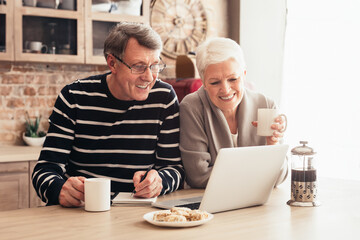 A happy senior couple enjoys a relaxing afternoon in their kitchen. The woman is holding a cup of coffee while looking at a laptop screen. Her husband is taking notes