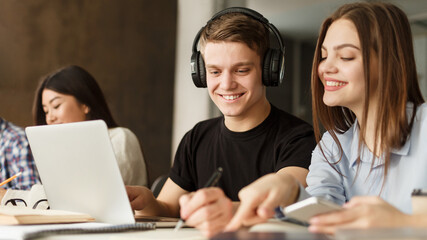 A cheerful young guy wearing headphones sits at a table with a laptop, deeply engaged in a study session alongside fellow students.