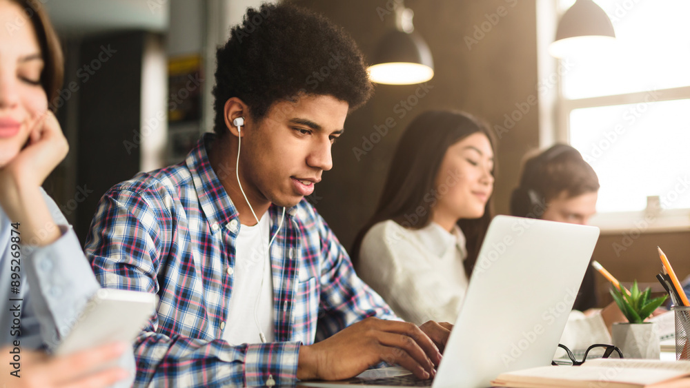 Poster a focused young black man with headphones works intently on a laptop in a bustling library environme
