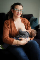 Fototapeta premium Young mother breastfeeding her baby, the woman is sitting on a sofa and smiling