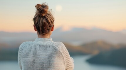 Fototapeta premium A woman with a top bun in a white sweater gazes over a scenic landscape with mountains and a body of water, symbolizing contemplation and serenity at sunset.