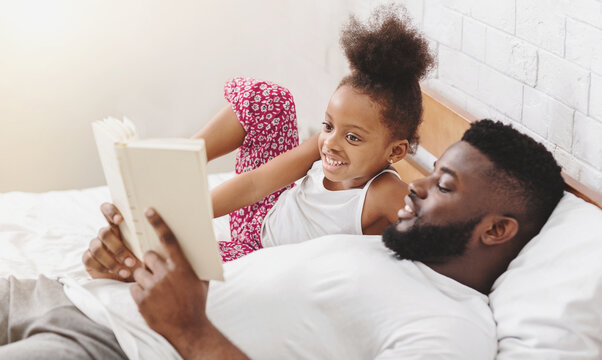 A father is lying in bed with his little daughter. He is reading a book to her. They both have smiles on their faces, lying on a white sheet and the headboard is white with a light grey accent.