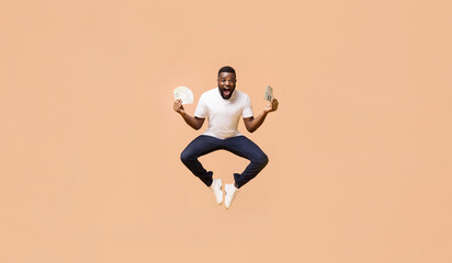 African American young man in a white t-shirt and dark jeans is jumping in the air, with his arms outstretched, holding stacks of money in each hand. The background is a soft peach color.