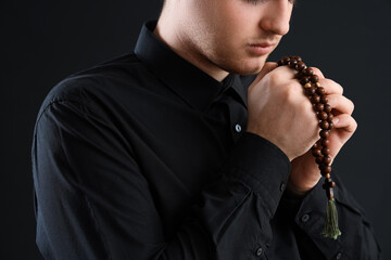Young man praying with beads on dark background, closeup