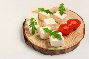 Wooden board with delicious feta cheese pieces, tomatoes and arugula on white grunge background