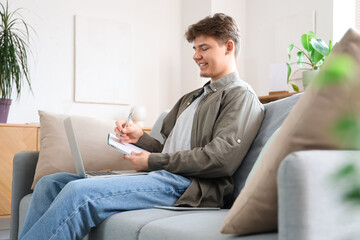 Male student with notebook and laptop studying online on sofa at home