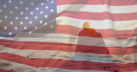 Image of american flag over happy diverse couple wrapped in rug embracing sat on sunset beach