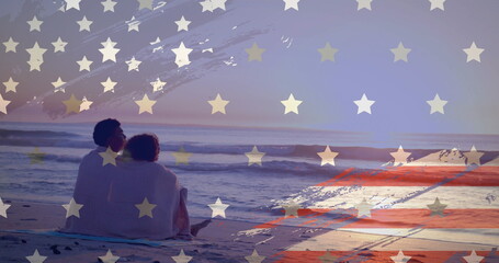 Image of american flag over happy diverse couple wrapped in rug embracing sat on sunset beach