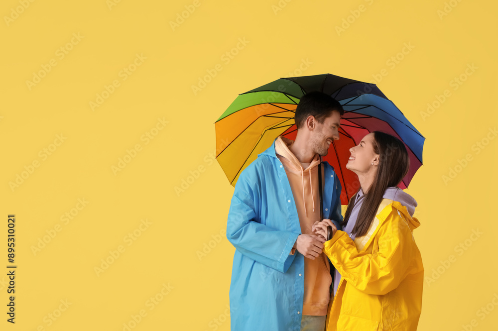 Wall mural Young couple in raincoats with umbrella on yellow background