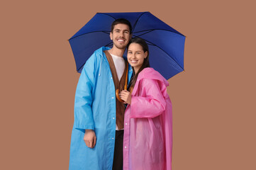 Young couple in raincoats with umbrella on brown background